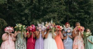 Bridesmaids in mixed coloured dresses hold coordinating bouquets in front of their faces.